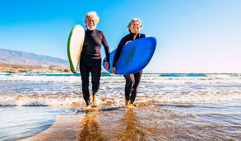 Retired couple walking through water with surfboards