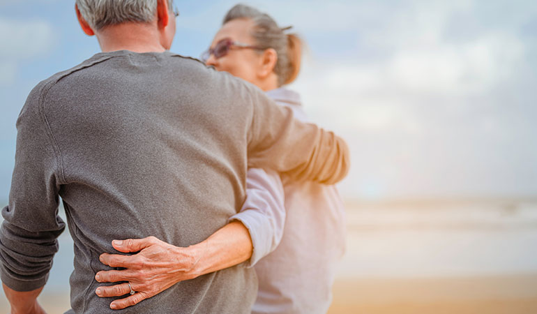 Retired couple sat on beach with arms around one another